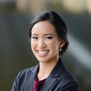 A headshot of Nicole Dailo. Nicole is an Asian woman with dark hair tied into a bun. She wears a pinstripe gray blazer and a maroon shirt.